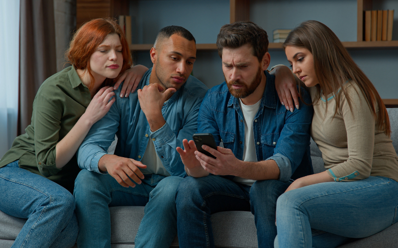 Impactos de um erro de compliance:Na imagem quatro jovens adultos sentados em um sofá, olhando com expressão séria e preocupada para o celular que um deles segura. Eles parecem concentrados ou impactados com o conteúdo exibido na tela. O ambiente é uma sala com prateleiras ao fundo.